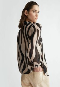 Blusa traslucida in beige con strisce astratte nere, caratterizzata da un design con colletto e maniche arricciate. Abbinata a pantaloni beige chiaro.
