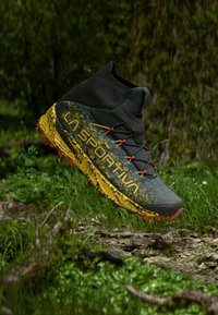 Svart og gul La Sportiva terrengløpesko i luften over en mudret, gressdekket skogbunn med små planter og steiner synlige.