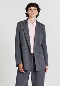 Persona con capelli ricci corti che indossa un blazer grigio a righe e pantaloni sopra una camicia a bottoni rosa chiaro, in piedi davanti a uno sfondo neutro.