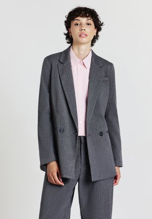 Persona con capelli ricci corti che indossa un blazer grigio a righe e pantaloni sopra una camicia a bottoni rosa chiaro, in piedi davanti a uno sfondo neutro.