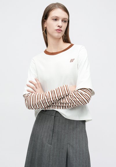 Άσπρη κοντομάνικη μπλούζα με καφέ ριγέ μακριά μανίκια, καφέ γιακά σε αντίθεση και λεπτομέρεια λογότυπου στο στήθος, συνδυασμένη με γκρι πλισέ φούστα.