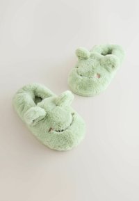 Zapatillas de felpa verdes con forma redondeada, textura suave y acentos de orejas. Presenta una cara sonriente con detalles en rosa.