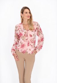 Blusa rosa a maniche lunghe con motivo floreale, scollatura a V, chiusura con bottoni frontale e dettagli arricciati alle spalle, abbinata a pantaloni aderenti beige.