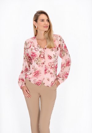 Blusa rosa a maniche lunghe con motivo floreale, scollatura a V, chiusura con bottoni frontale e dettagli arricciati alle spalle, abbinata a pantaloni aderenti beige.