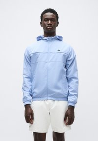 Casaco azul claro com fecho de correr, capuz, punhos elásticos e um detalhe subtil de costura horizontal. Apresenta um logótipo verde no peito.