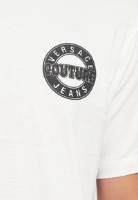 Baltas medvilninis marškinėlių suapvalintu juodu logotipu, kuriame yra tekstai "VERSACE" ir "COUTURE JEANS". Minkšta tekstūra, klasikinis siluetas, minimalistiškas dizainas.