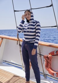 Navy jaquard-huppari valkoisin raidoin, jossa on etutasku ja nauhat. Yhdistettynä tummansinisiin verkkareihin. Taustalla puinen terassi ja meri.