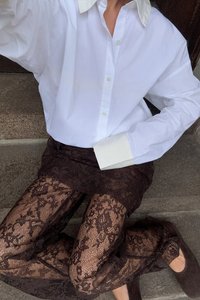Camicia bianca oversize con colletto leggero, abbinata a leggings di pizzo marrone trasparente. Tacchi chiusi di colore marrone completano l'outfit.
