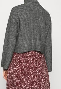 Jersey gris recortado con cuello alto y mangas anchas, combinado con un vestido de estampado floral burdeos con pequeños acentos en blanco.