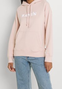 Ljusrosa hoodie gjord av mjukt tyg, med en framficka, snörning på huvan och vit Levi's-logotyp tryckt på bröstet.