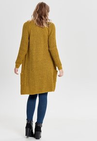 Hořčicově žlutý pletený cardigan s žebrovanou texturou, dlouhými rukávy a dvěma předními kapsami. Stylizováno s modrými džínami a kotníkovými botami.