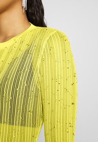 Gul meshtröja med vertikala ränder, dekorerad med spridda paljetter för textur. Inkluderar rund halsringning och långa ärmar.