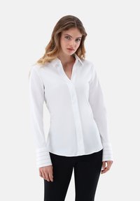 Camicia bianca a maniche lunghe con scollo a V e polsini a strisce. Texture liscia, vestibilità su misura e un leggero splendore completano il design pulito.