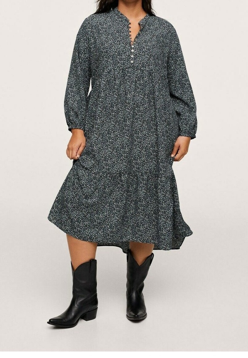 Femme portant une robe à manches longues, aux genoux, sombre et à motifs, et des bottes de cowboy noires mi-mollet, debout devant un fond uni.