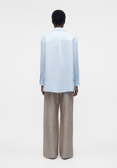 Άτομο με κοντά σγουρά μαλλιά που φορά χαλαρό γαλάζιο πουκάμισο με μακριά μανίκια και φαρδιά μπεζ παντελόνια με ρίγες, στέκεται με την πλάτη προς τα εμπρός.