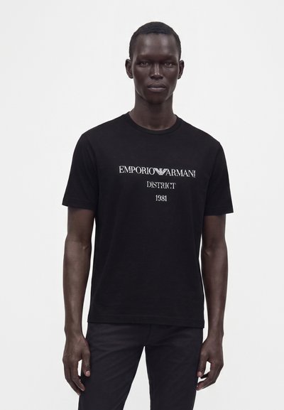 Άνδρας που φοράει μαύρο t-shirt Emporio Armani με άσπρο κείμενο "District 1981", στέκεται μπροστά σε απλό λευκό φόντο.