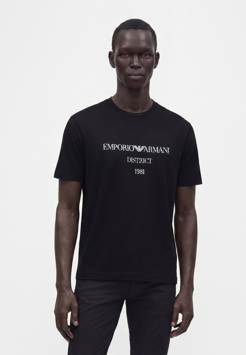 Άνδρας που φοράει μαύρο t-shirt Emporio Armani με άσπρο κείμενο "District 1981", στέκεται μπροστά σε απλό λευκό φόντο.