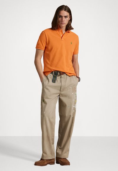 Oranje polo-shirt met een groen logo, gecombineerd met beige broeken met verfspetters. Bruine schoenen maken de outfit compleet.
