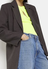 Tmavě šedý blazer s volnějším střihem, spárovaný s jasně žlutým žebrovaným topem s výřezem. Viditelné světle modré džíny s vysokým pasem.