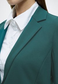 Blazer bleu sarcelle avec revers structurés, associé à une chemise blanche à boutons. Le blazer présente une texture lisse et une coupe ajustée.