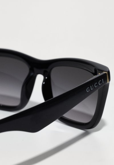 Juodos saulės akinių su gradientiniais pilkais objektyvais. Storas plastikinis rėmas su lygia tekstūra. Rėmuose yra auksinė detalė, ant kurios užrašyta "GUCCI".