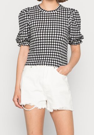Frau trägt ein schwarz-weiß kariertes kurzes Oberteil mit Puffärmeln und weiße, ausgefranste Jeansshorts, eine Hand in der Tasche.