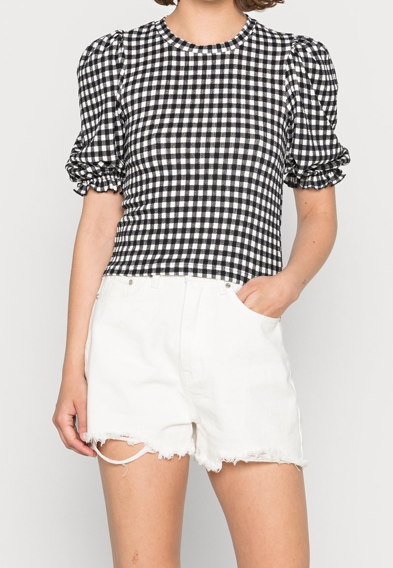 Femme portant un haut court à manches bouffantes à carreaux noir et blanc avec un short en jean blanc effiloché, une main dans la poche.