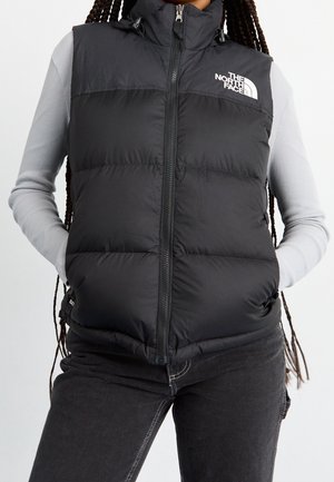 Osoba nosząca czarną pikowaną kamizelkę North Face na jasnoszarej, prążkowanej bluzce z długim rękawem i ciemnych dżinsach, z rękami w kieszeniach kamizelki.