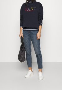 Felpa blu marina con logo multicolore "GANT", rifiniture bianche, abbinata a jeans di denim blu e sneakers bianche. Una borsa nera è tenuta in mano.