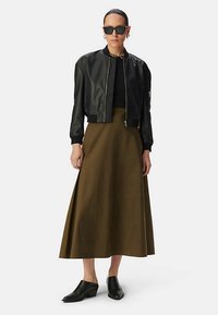 Zwarte leren bomberjack, zwart topje, olijfgroene midi-rok, zwarte schoenen met blokhak. Vrouw draagt oversized zonnebril en oorbellen.