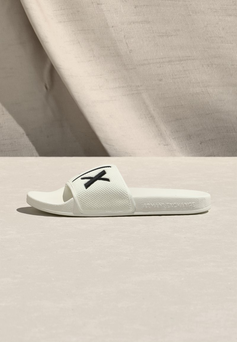 Weiße Slide-Sandale mit einem strukturierten Riemen, der ein schwarzes Logo zeigt. Hergestellt aus synthetischem Material, hat sie ein flaches, offen Zehen-Design.