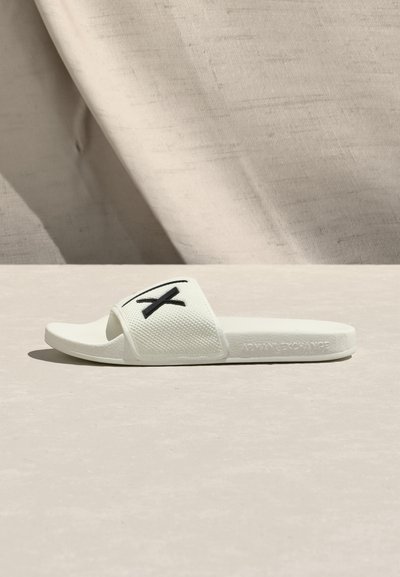 Sandale blanche à semelle plate avec une bride texturée portant un logo noir. Fabriquée en matériau synthétique, elle présente un design ouvert aux orteils.
