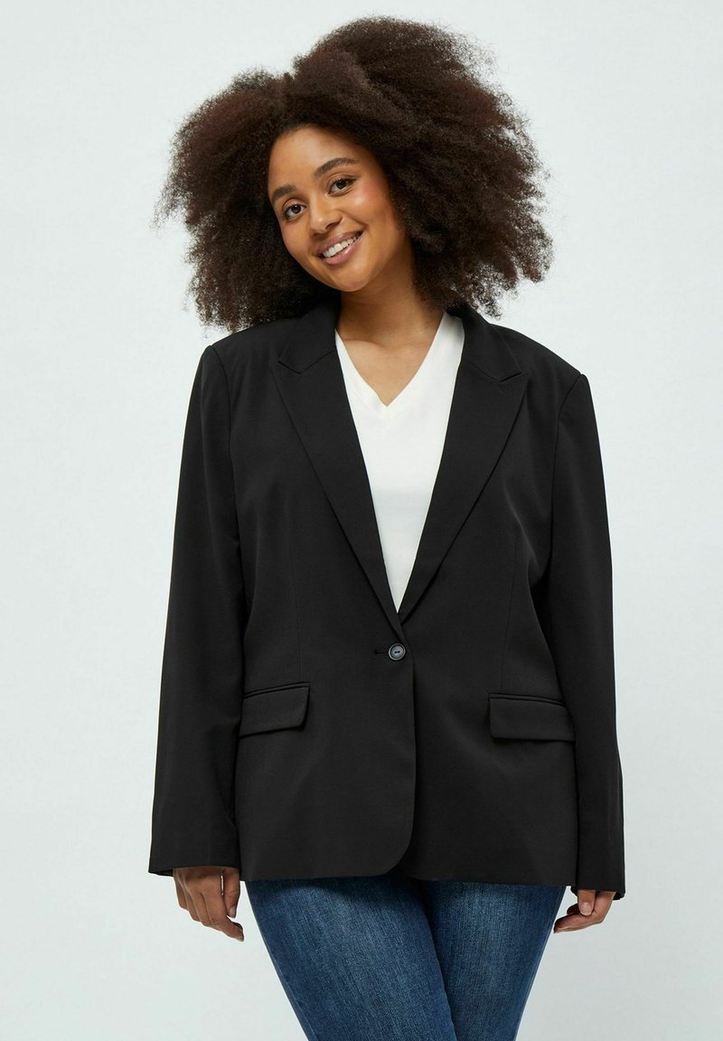 Blazer nero con revers a punta, chiusura con un solo bottone, due tasche frontali e una vestibilità sartoriale. Indossato sopra un top bianco a V.