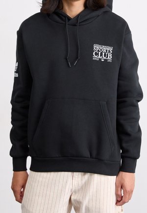 Osoba ubrana w czarną bluzę z kapturem Adidas Originals Sports Club z przednią kieszenią i białe spodnie w paski, stojąca na jednolitym tle.