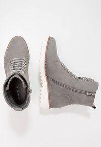 Bottes à chevilles en daim gris avec bout rond, semelle blanche en caoutchouc, devant à lacets, fermeture éclair latérale et détail de couture contrastante marron.