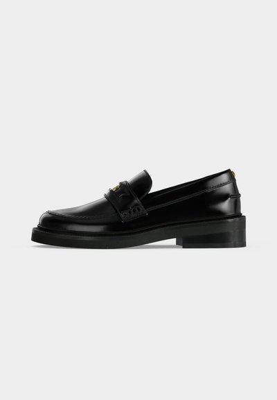 Mocassin en cuir noir poli avec un petit talon carré, surpiqûres moc toe et une bride décorative sur le dessus.