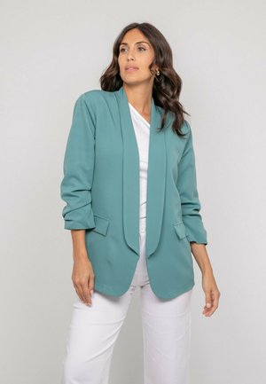 Femme aux cheveux ondulés et foncés, portant un blazer turquoise aux manches retroussées, un haut blanc et un pantalon blanc, regardant sur le côté sur un fond uni.