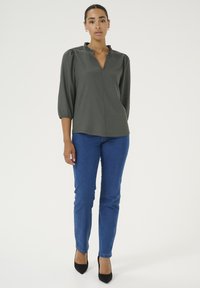 Grüne strukturierte Bluse mit Rüsche am Kragen, 3/4-Ärmeln, kombiniert mit blauen enger geschnittenen Jeans und schwarzen spitzen Absatzschuhen.
