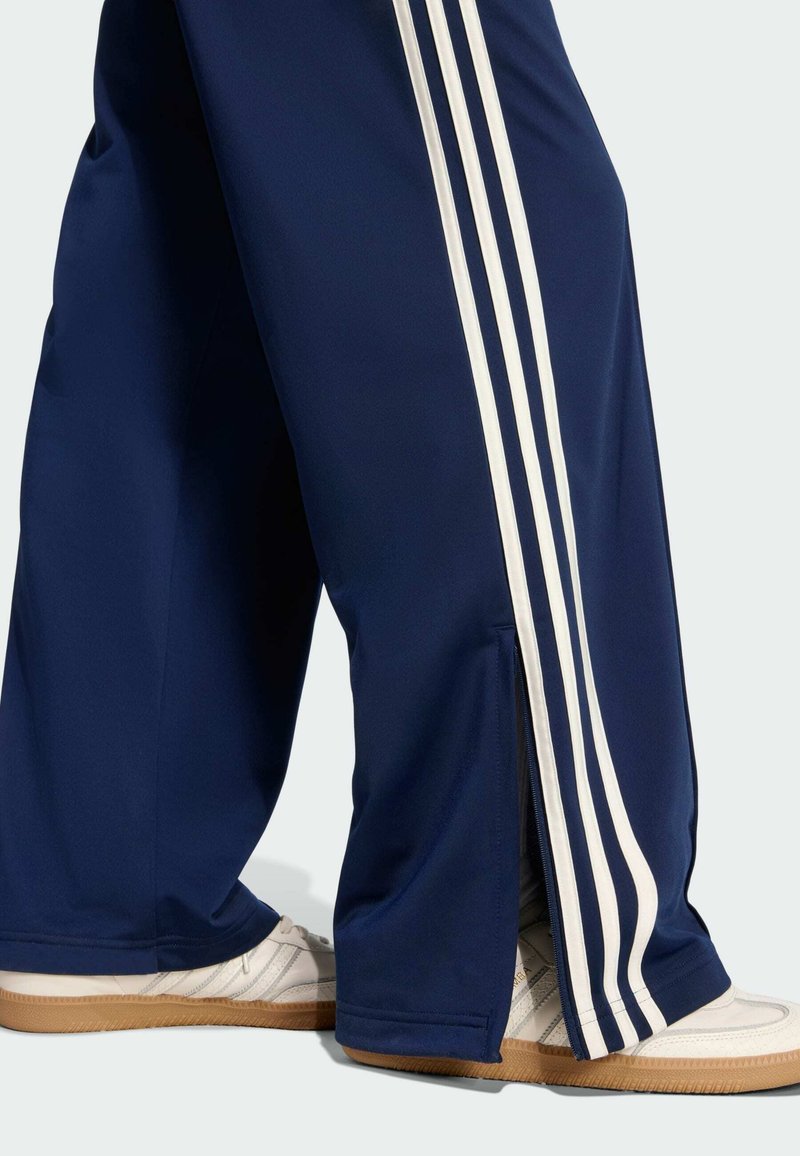 Pantalon de sport bleu marine avec des bandes blanches sur les côtés, doté d'une coupe décontractée et d'ouvertures zippées aux chevilles. Porté avec des chaussures blanches.