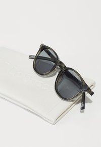 Ochelari de soare cu lentile gri închis, ramă transparentă închisă și brațe drepte, așezați pe un capac alb din piele cu inscripția "Le Specs."
