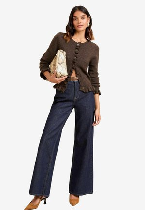 Cardigan en laine marron avec des bords froncés, boutonné à l'avant, porté sur un jean flare en denim foncé taille haute. Tenant un sac en simili serpent beige.