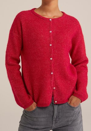 Personne portant un cardigan en tricot rouge boutonné avec des manches longues et les mains dans les poches d’un pantalon gris sur un fond uni.