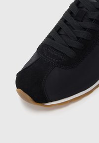 Μαύρο αθλητικό παπούτσι με συνδυασμό σουέτ και συνθετικών υλικών. Στρογγυλεμένη μύτη, χοντρές μαύρες κορδόνια και εξωτερική σόλα από καουτσούκ χρώματος καραμέλας για καλύτερη πρόσφυση.