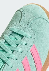 Baskets en suede vert avec des rayures roses en zigzag, lacets ronds et semelle en caoutchouc gomme. La vue rapprochée met en valeur la texture et les détails de couture.