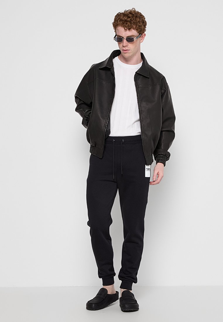 Giubbotto bomber in pelle nera, maglietta a coste bianca, pantaloni della tuta neri con cintura elastica e scarpe slip-on nere. Il modello indossa occhiali da sole.
