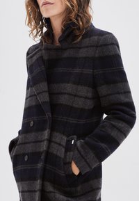 Woman wearing a dark blue and gray horizontal striped wool coat, hands in pockets, standing against a plain light background.