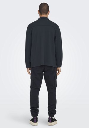 Mann steht mit dem Rücken zur Kamera, trägt ein schwarzes Langarmshirt, schwarze Cargohosen und schwarze Sneakers mit weißen Sohlen und lila Akzenten.