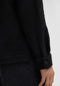 Camicia nera con texture, dal taglio rilassato, con polsini abbottonati e dettagli di cucitura visibili. Il materiale sembra morbido e leggermente peloso.