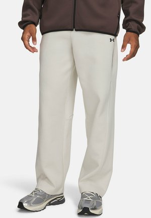Man draagt beige Under Armour broek, zilveren en zwarte hardloopschoenen en een bruine jas met rits, met ontspannen handen langs de zijkant.