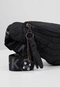 Sac banane noir matelassé avec fermeture éclair, bandoulière réglable avec inscription "NA-KD" et petit porte-clés parapluie avec étiquette circulaire attachée.